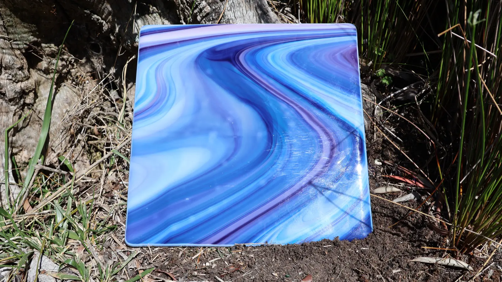 Large square fused-glass plate of flowing cobalt, periwinkle, and violet agate streaks, leaning against a weathered tree trunk beside green rushes